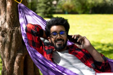 Memnun Hindu genç adam telefonda konuşuyor ve hamakta yatıyor.