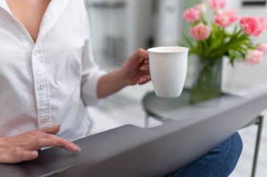 Bilinmeyen bir kadın elinde kahve bardağıyla ev bilgisayarında çalışıyor..