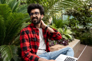 Kırmızı kareli gömlekli adam dizüstü bilgisayarda çalışıyor ve yorgun görünüyor.