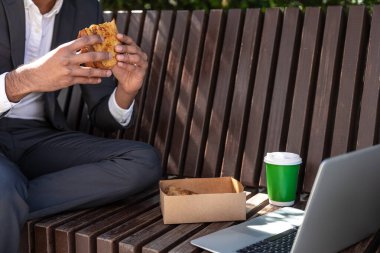 Genç adam dışarıda bankta otururken yemek yiyor ve dizüstü bilgisayarla çalışıyor.