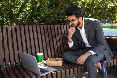 Genç adam dışarıda bankta otururken yemek yiyor ve dizüstü bilgisayarla çalışıyor.