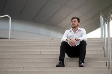 Kendine güvenen bir adam. Beyaz gömlekli kendine güvenen genç adam merdivenlerde oturuyor.