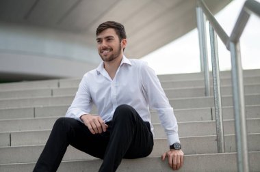 Kendine güvenen bir adam. Beyaz gömlekli kendine güvenen genç adam merdivenlerde oturuyor.