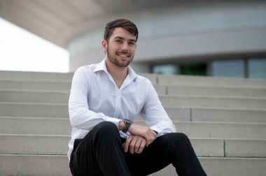 Kendine güvenen bir adam. Beyaz gömlekli kendine güvenen genç adam merdivenlerde oturuyor.