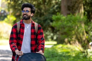 Genç baba. Ekose gömlekli genç adam parkta bebek arabası taşıyor.