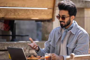 Bir kafede. Koyu saçlı Hintli adam pizza yiyor ve dizüstü bilgisayarla çalışıyor.