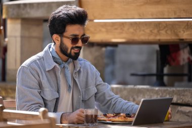 Bir kafede. Koyu saçlı Hintli adam pizza yiyor ve dizüstü bilgisayarla çalışıyor.