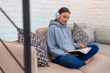 Kulaklıkla müzik dinleyen ve kitap okuyan bir kadın evdeki kanepede otururken..