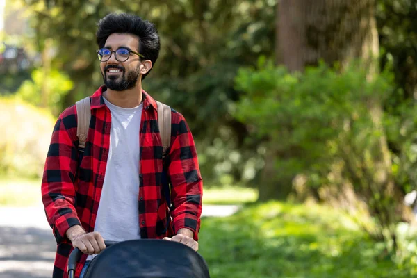 Genç baba. Ekose gömlekli genç adam parkta bebek arabası taşıyor.