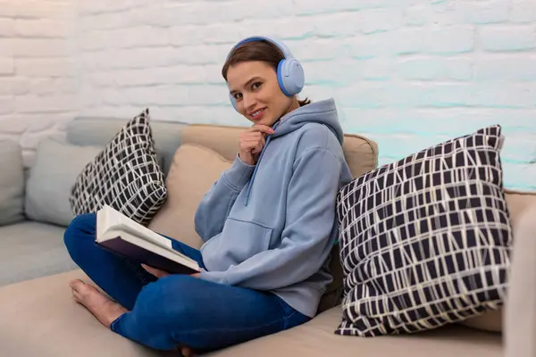 Oturma odasında kanepede otururken kulaklıklardan müzik dinleyen ve kitap okuyan genç bir kadın..