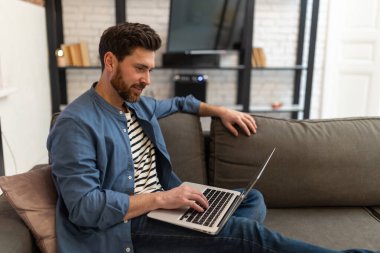 Genç sakallı yakışıklı adam evde oturuyor ve dizüstü bilgisayarla çalışıyor.