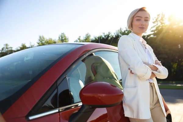 Attractive woman in formal wear standing near red car on parking before business trip