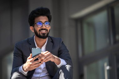 Koyu renk saçlı Hintli adam merdivenlerde elinde telefonla oturuyor.