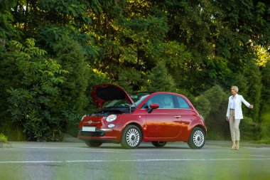 Yetişkin bir kadın otostop çekerken kırmızı arabanın yanında duruyor ve şoförlerden yardım istiyor.