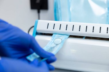 Tanınmayan doktor sterilizasyon prosedürü uyguluyor. Ellerinde mavi eldivenlerle dişçi aletleri tutuyor.