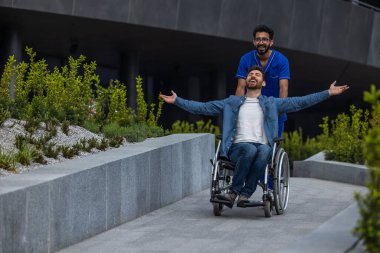 Bir hastane bahçesinde. Tekerlekli sandalyedeki adam erkek hemşireyle yürüyor.