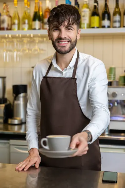 カフェでコーヒーカップを保持するエプロンを着用した笑顔のひげ付き男 — ストック写真