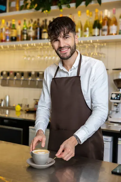 カフェでコーヒーカップを開催するエプロンを着た陽気な男バリスタは カフェの訪問者に楽しい飲み物を提供します — ストック写真