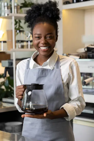 カフェカウンターの近くに立っている醸造コーヒーとガラス鍋を保持する陽気な多文化女性ウェイター — ストック写真
