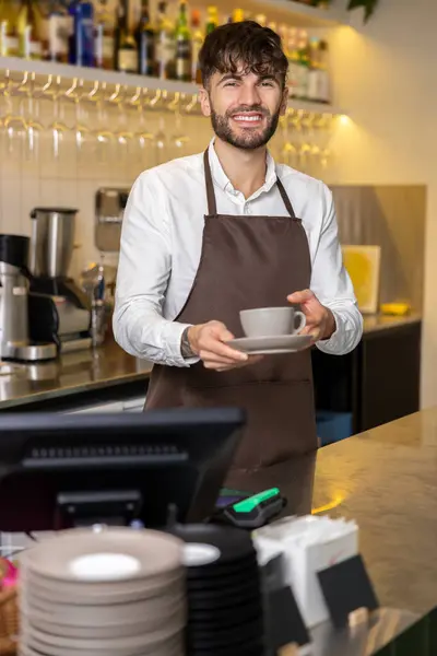 カフェでコーヒーカップを保持するエプロンを着用したベアードマンは カフェパトロンのおいしい飲み物を作るバリスタとして働きます — ストック写真