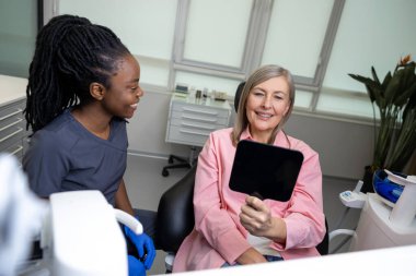 Beyaz kadın hasta dişçinin ofisinde aynaya bakıp gülümsüyor.