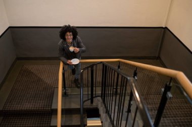 Kahve içiyorum. Merdivenlerde elinde bardakla gözlüklü, kıvırcık saçlı genç adam.