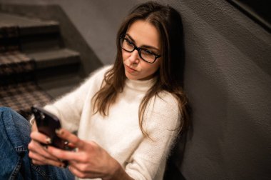 Merdivenlerde dikilen sıradan elbiseli genç bir kadın oteldeki boş zamanlarında cep telefonuyla internette geziniyor.