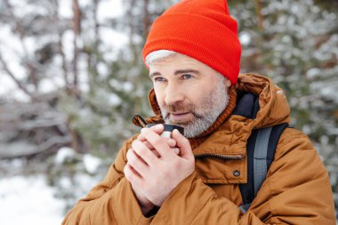 Soğuk bir gün. Sakallı, kır saçlı, kırmızı şapkalı, elinde termosla bir kış ormanında