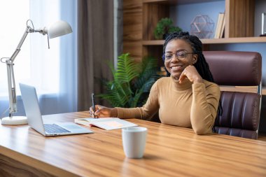Rasta bukleli neşeli kadın ev ofisinin iç kısımlarında uzaktan bilgisayar ve teknoloji kullanıyor.