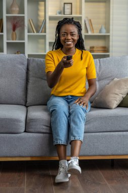 Sevimli esmer tenli genç kadın televizyon seyrediyor ve kontrol panelini elinde tutuyor.