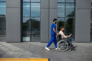 Hastayla yürü. Erkek hemşire tekerlekli sandalyede sakat bir hastayı yürüyüşe çıkarıyor.