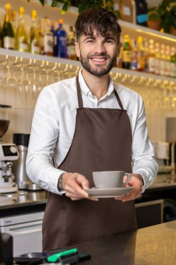 Kahve dükkanının tezgahında duran sakallı barista. Müşteri için bir fincan kahve tutuyor.