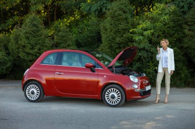 Telefonla konuşan bir kadın yolda bozuk arabasının yanında yardım istiyor. Arka planda yeşil orman var.