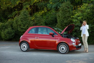 Genç bir kadın yolda dikilirken arabasını tamir etmek için cep telefonu kullanıyor.