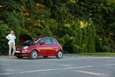 Telefonla konuşan bir kadın yolda bozuk arabasının yanında yardım istiyor. Arka planda yeşil orman var.