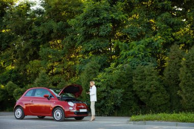Genç bir kadın yolda dikilirken arabasını tamir etmek için cep telefonu kullanıyor.