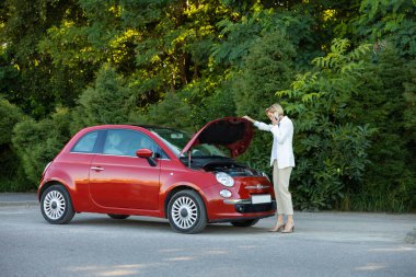 Telefonla konuşan bir kadın yolda bozuk arabasının yanında yardım istiyor. Arka planda yeşil orman var.