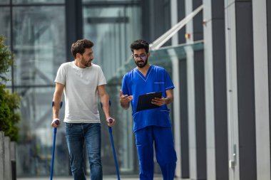 Rehabilitasyondaki adam. Koltuk değnekleriyle yürüyen ve erkek hemşireyle konuşan genç sakallı adam.