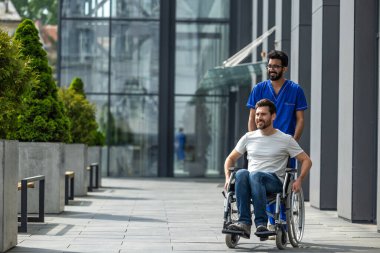 Engelli. Esmer erkek hemşire bir hastayla tekerlekli sandalyeyi yuvarlıyor.