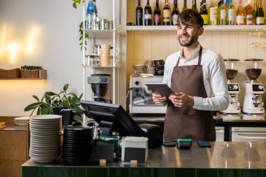 Barda ya da kafede tablet bilgisayarı olan önlüklü mutlu bir adam ya da garson.