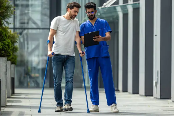 Man Rehabilitation Young Bearded Man Crutches Walking Talking Male Nurse — Stock Photo, Image