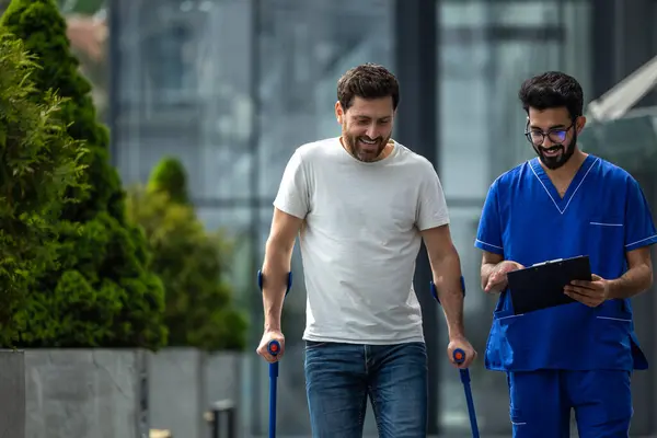 Walk together. Man with crutches and a male nurse talking friendly on a walk