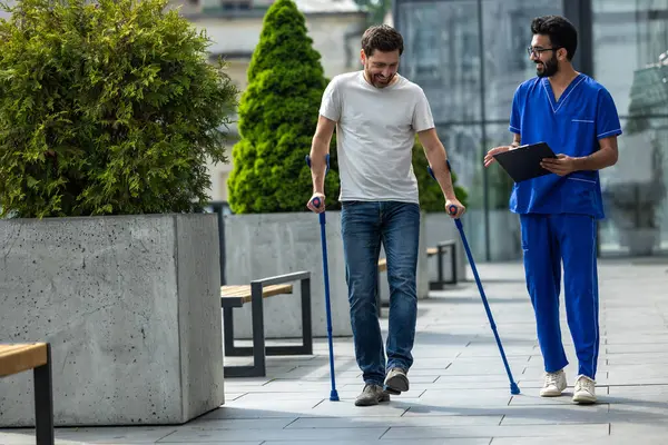 Walk together. Man with crutches and a male nurse talking friendly on a walk