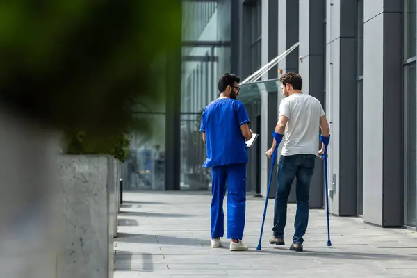 Rehabilitation. Man with crutches and a male nurse in a blue uniform walking in a hospital yard