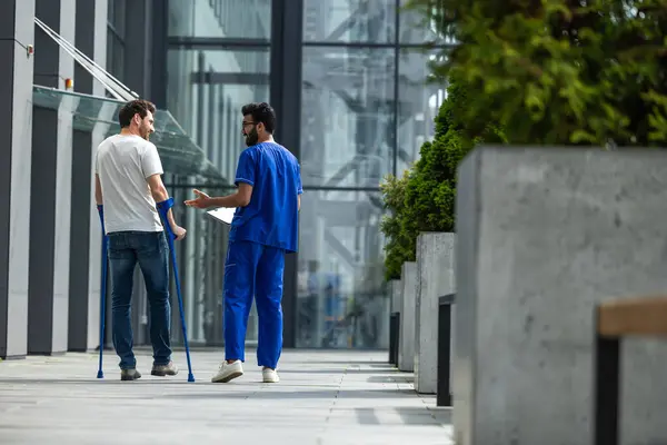 Rehabilitation. Man with crutches and a male nurse in a blue uniform walking in a hospital yard