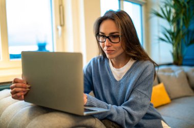 Gözlüklü güzel beyaz kadın otel odasında bilgisayarını kullanarak uzaktan kumandasını yapıyor.