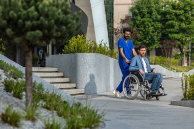 Hastayla yürü. Mavi doktorlu erkek hemşire tekerlekli sandalye taşıyor.