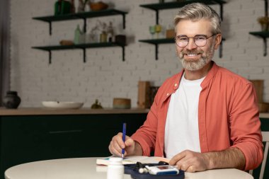 Glukometreyle gülen gri saçlı orta yaşlı adam kan şekeri seviyesini kontrol ediyor. Not defterinde göstergelerle birlikte not alıyor.