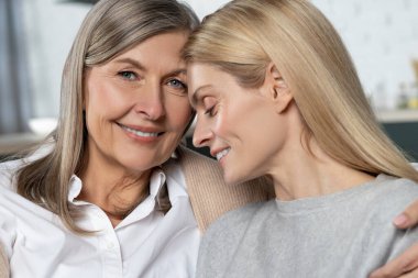 Şirin yaşlı kadın ve yetişkin kızı evdeki kanepede otururken sarılıp gülümsüyorlar.