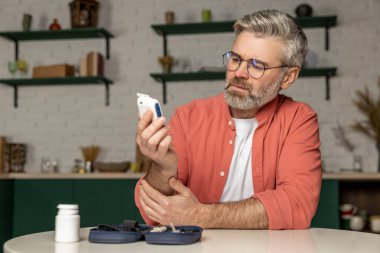 Glukometresi olan gri saçlı diyabetik adam evde kan şekerini ölçüyor.